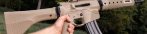 Close-up of a person holding a tan AR-style rifle outdoors with trees in the background